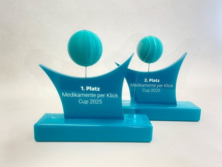 Award für den Medikamente Per Click Cup 2025 aus türkisem Acrylglas mit Lasergravur. Die Gravur wurde mit weißem Lack ausgelegt. Die Köpfe der stilisierten Figuren sind aus 3D-gedrucktem PETG Material und sehen aus wie Basketbälle.