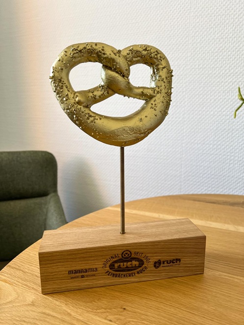 Award aus lasergraviertem Eichenblock mit Gewindestange, auf der eine goldene Brezel platziert ist. Der Award ist für eine Bäckerei-Kette.