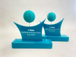 Award für den Medikamente Per Click Cup 2025 aus türkisem Acrylglas mit Lasergravur. Die Gravur wurde mit weißem Lack ausgelegt. Die Köpfe der stilisierten Figuren sind aus 3D-gedrucktem PETG Material und sehen aus wie Basketbälle.