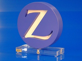 Foto zeigt Award aus Acrylglas. Der Award ist rund, in der Mitte ist ein verschnörkeltes Z. Der runde Kreis ist dunkelblau lackiert, darin ist ein weißes Z. Der Kreis ist auf einer Grundplatte aus massiven, farblosem Acrylglas verschraubt.
