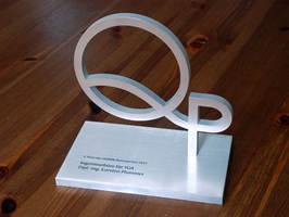 Aluminium Grundplatte mit farbig ausgelegter Stichelgravur, senkrecht aufgeschraubter Aluminium Schriftzug QP. Award steht auf einem dunklen Holztisch.