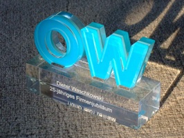 Foto zeigt Award mit großen Buchstaben OW aus Sandwich-Acrylglas (farblos / türkis / farblos), die auf einem massiven, farblosen Sockel verklebt sind. Auf dem Sockel ist eine Lasergravur für den Empfänger.
