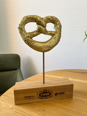 Award aus lasergraviertem Eichenblock mit Gewindestange, auf der eine goldene Brezel platziert ist. Der Award ist für eine Bäckerei-Kette.