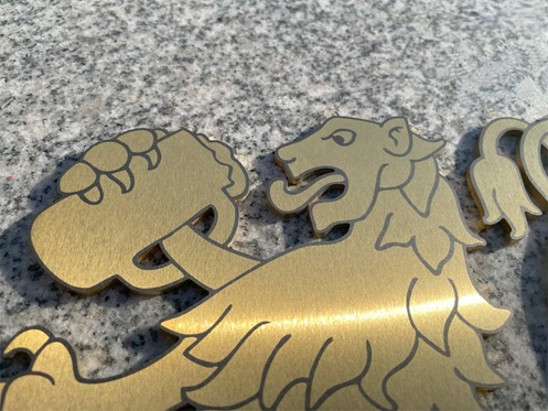 Foto zeigt einen Edelstahl-Laserzuschnitt mit einem heraldisch-anmutenden Löwen, der einen Bierkrug hält. Der Zuschnitt wurde TiN-Gold beschichtet und anschließend laserbeschriftet. Der Zuschnitt liegt auf einer Granitplatte.